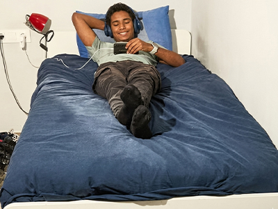 Joven con auriculares usando su celular mientras descansa en una cama azul.