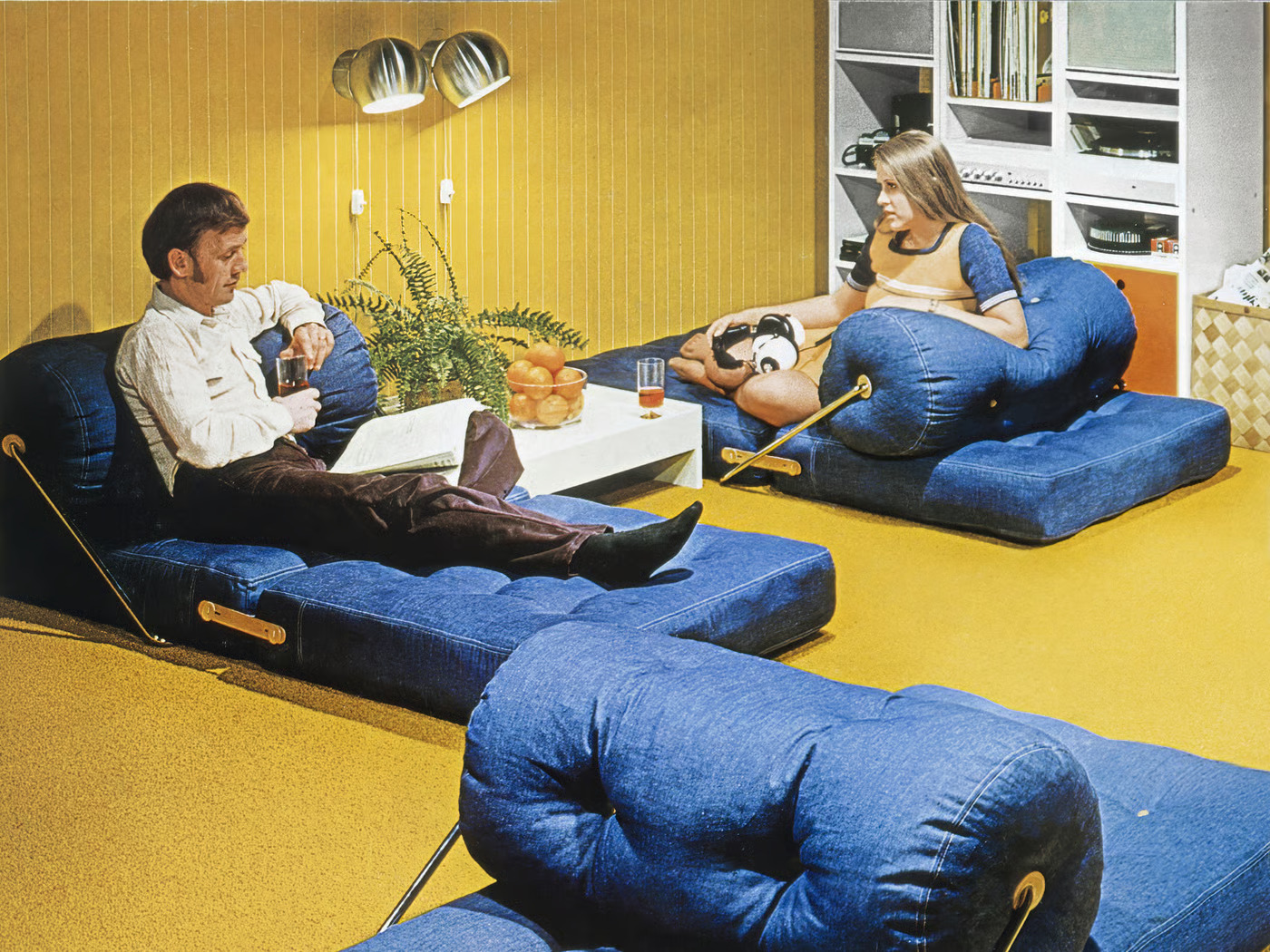 Pareja de los 70 en sillones de mezclilla, pared amarilla y muebles de audio modulares.