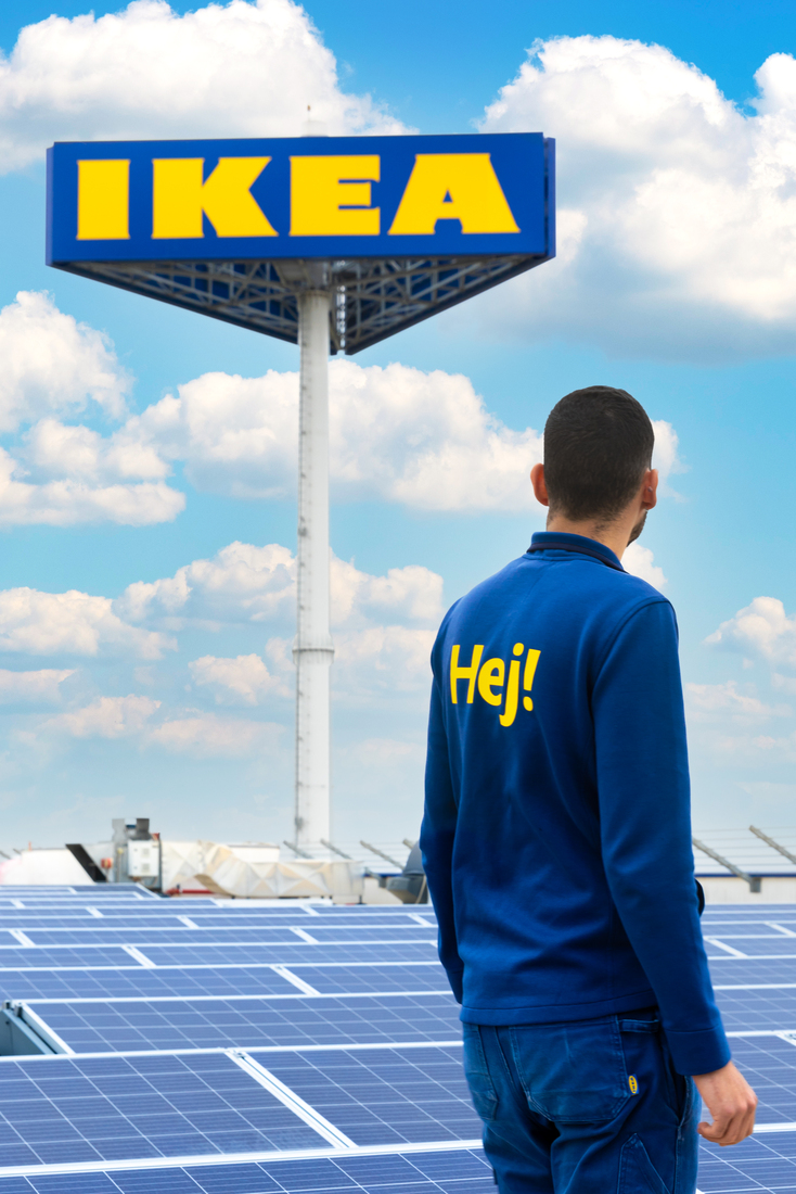 Un hombre con un uniforme de IKEA que dice "Hej!" de pie en un techo cubierto de paneles solares mirando hacia un letrero de IKEA.