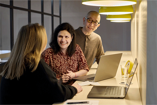 Three coworkers smiling and talking in front of their laptops at an office desk.
