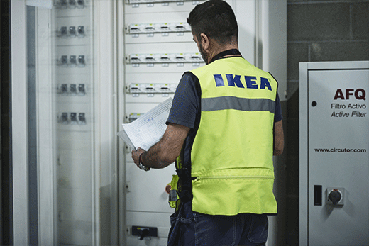 IKEA technician in a safety vest holding diagrams while inspecting an electrical panel.