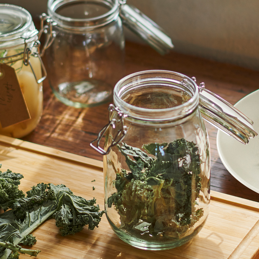 In a kitchen, a KORKEN jar with lid filled with kale chips is on a cutting board. Bits of kale are on the board near the jar.