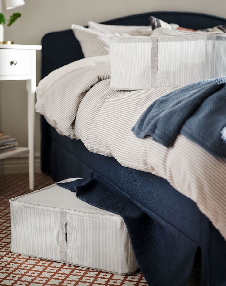 The side of a RAMNEFJÄLL bed with a throw and a HEMMAFIXARE storage case on top. Another storage case is under the bed.