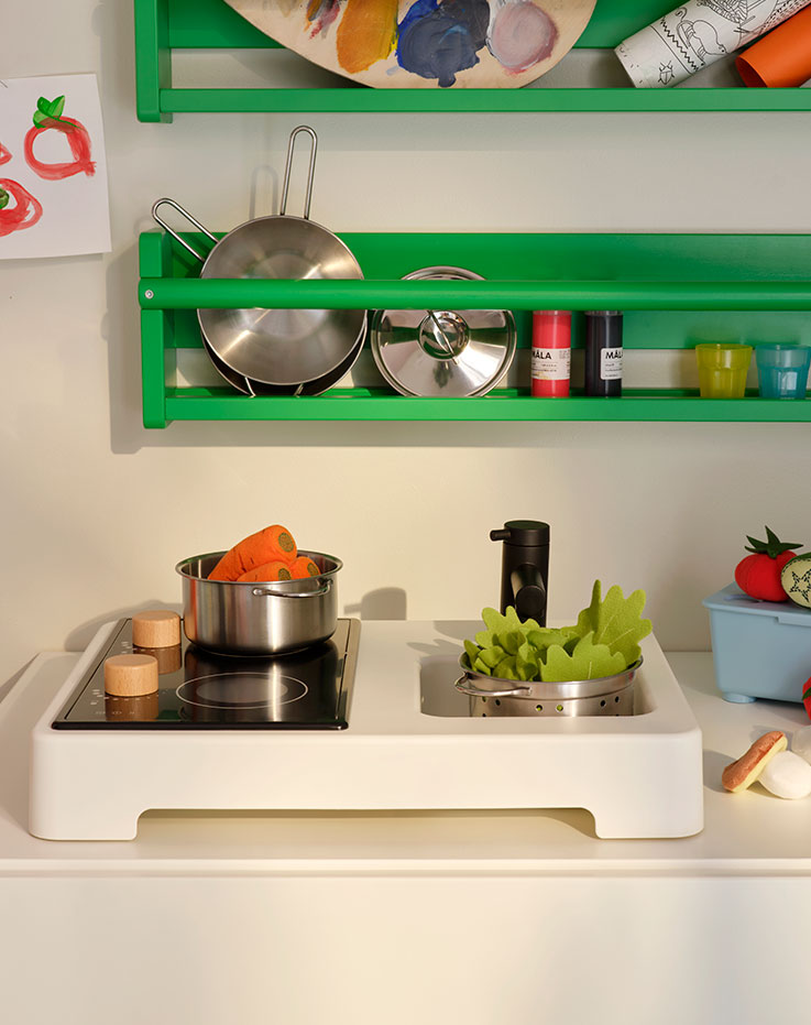 Parts of a STENMUS mini-play kitchen beneath green FLISAT wall storage, all filled with play-cooking accessories.