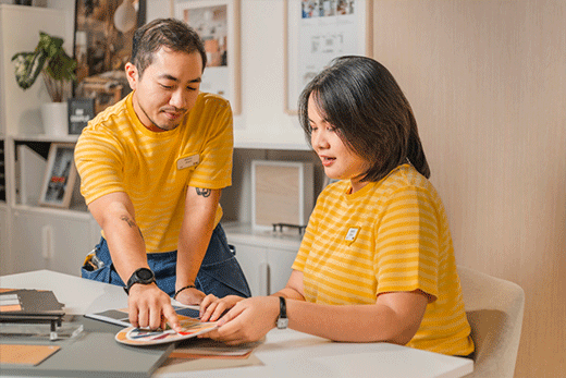 An IKEA man and woman in yellow striped t-shirts are standing and sitting around a table looking at samples.
