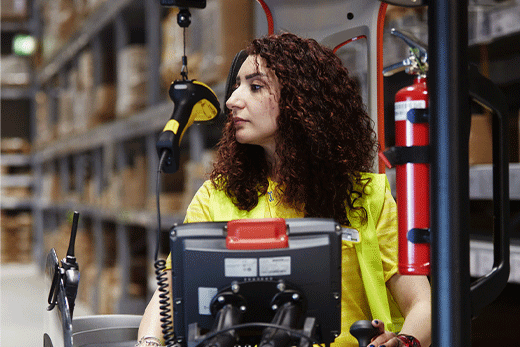 Woman in a safety vest operating a forklift in a warehouse with a handheld scanner and display.