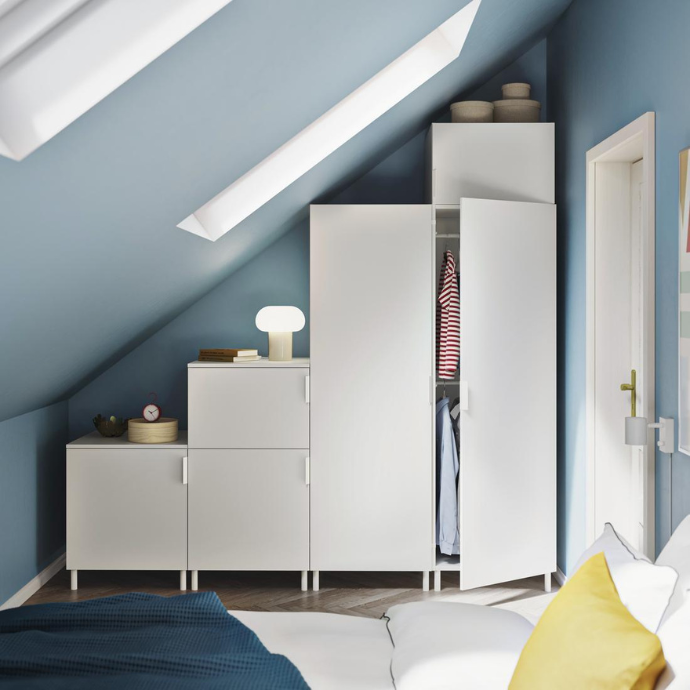 Modular white wardrobes of varying heights fitted under a sloped blue ceiling in a bright bedroom.