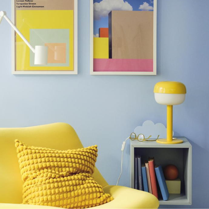 A cozy corner with a yellow armchair and textured pillow, featuring a yellow table lamp on a grey bedside shelf against a light blue wall with colorful framed art.