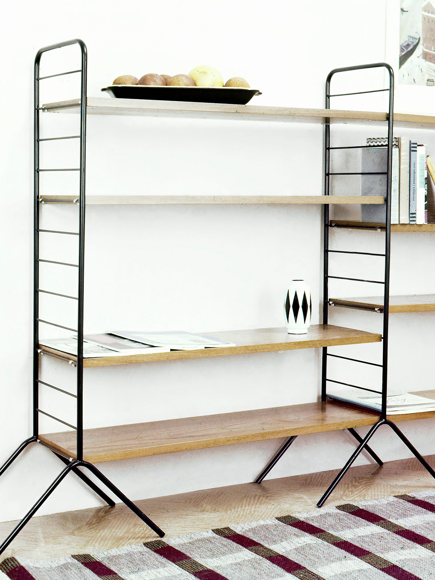 Minimalist modular shelving unit with a black metal frame and light wood shelves, decorated with books and a geometric vase.