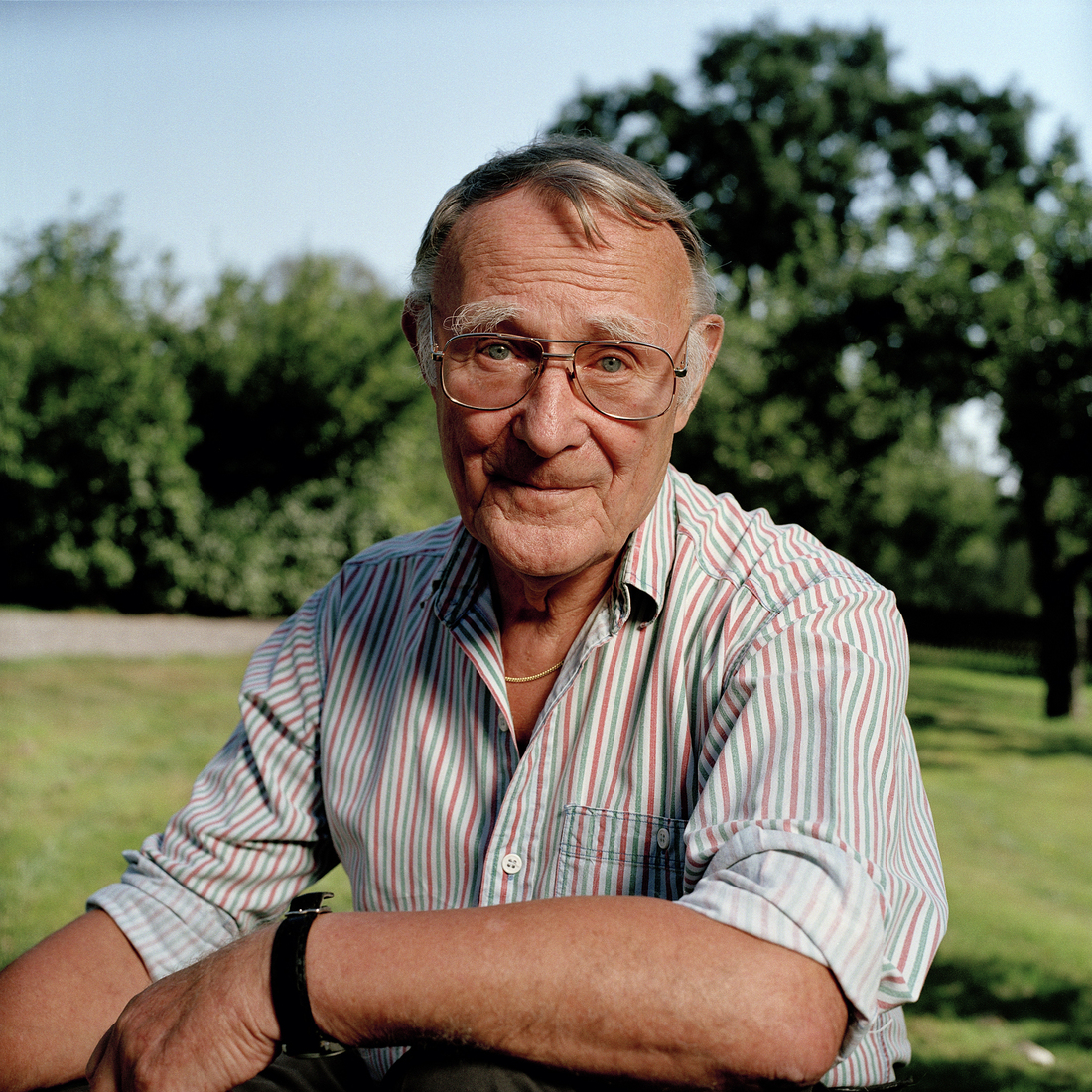 A close-up portrait of an elderly man wearing glasses and a striped shirt, smiling gently outdoors with trees in the background.