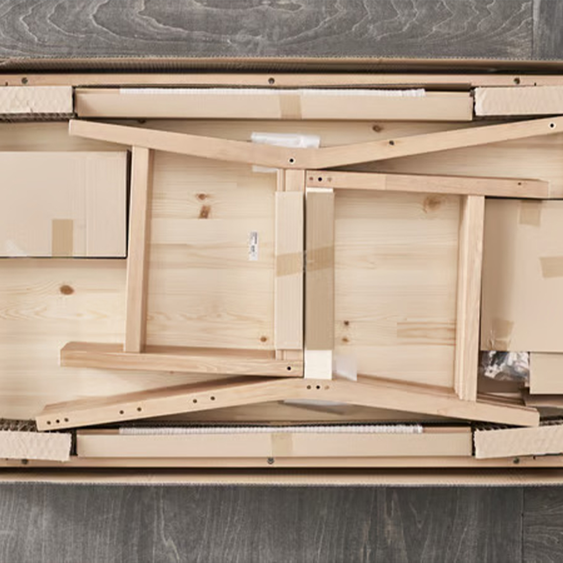 Overhead view of disassembled light wood furniture components neatly packed inside a flat cardboard shipping box.