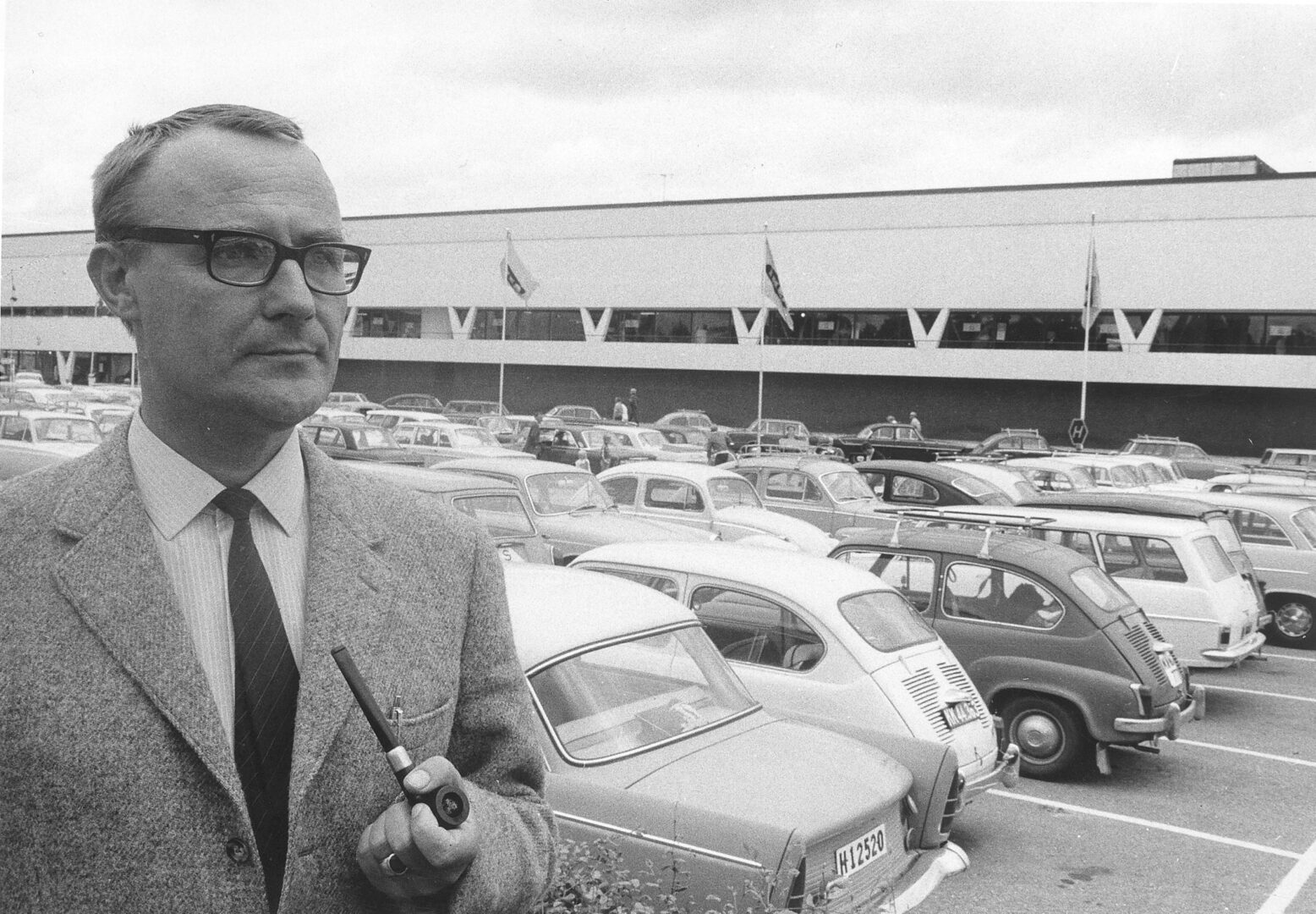 Foto antigua en blanco y negro de un hombre de traje con una pipa, con un estacionamiento lleno de autos clásicos y un edificio alargado al fondo.