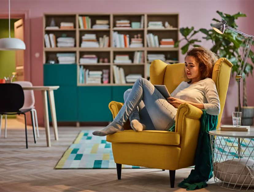 Una mujer relajada en un sillón orejero de color amarillo vibrante usando una tablet en una sala de estar moderna y colorida.