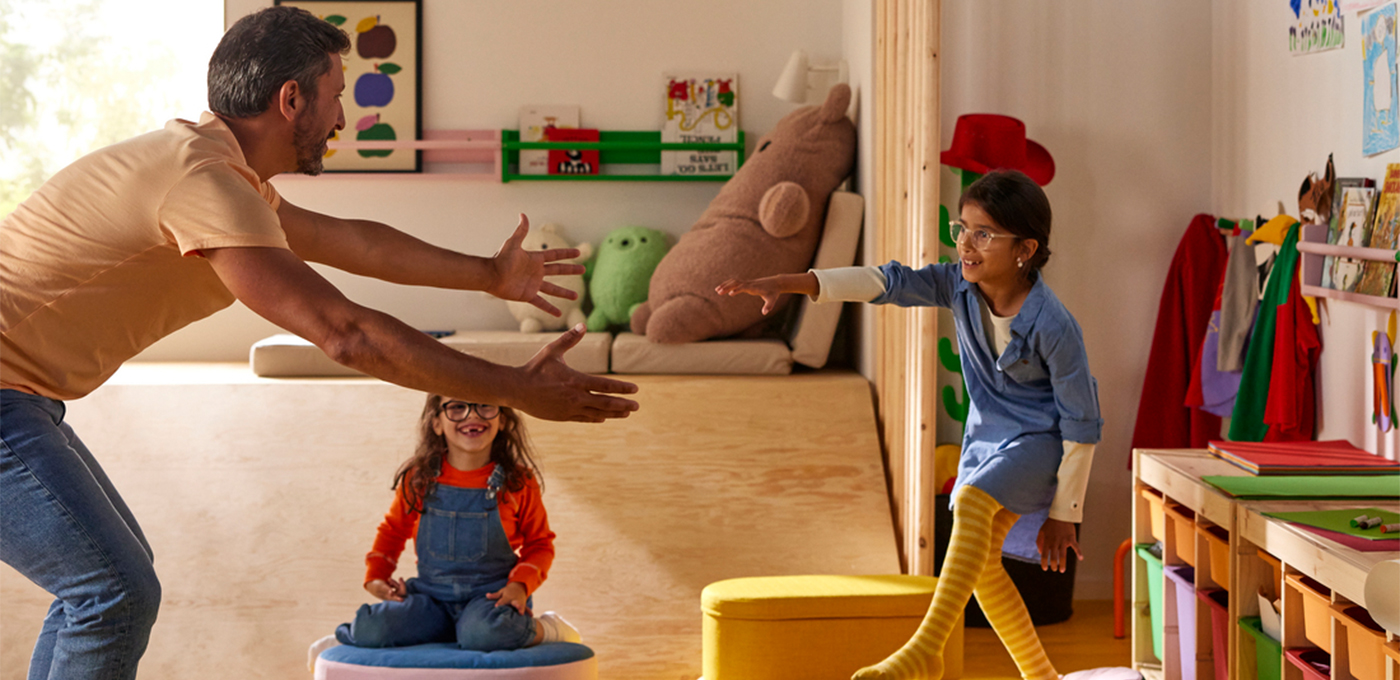 A father with open arms waits to hug his daughter in a colorful playroom, while another young girl sits nearby and watches with a smile.