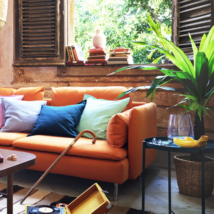 An orange sofa with colorful pillows in a rustic room, featuring a wooden cane leaning against it, a record player on the floor, and a large green plant next to a window with open shutters.