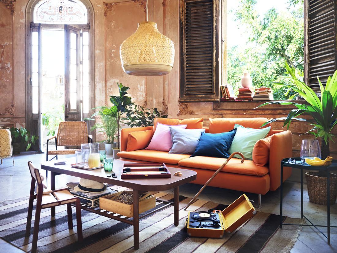 A vibrant living room with a bright orange sofa, colorful pillows, a wooden coffee table with games, and a woven pendant lamp, set in an airy room with rustic walls and open wooden shutters.